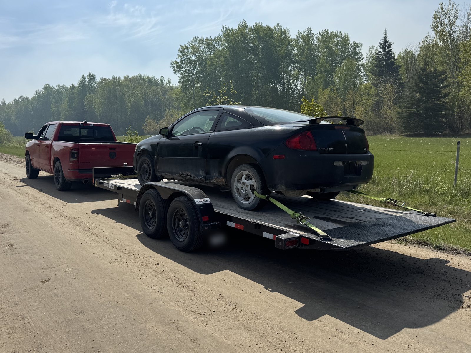 Coupe strapped to a tilt trailer behind a pickup truck on a gravel road
